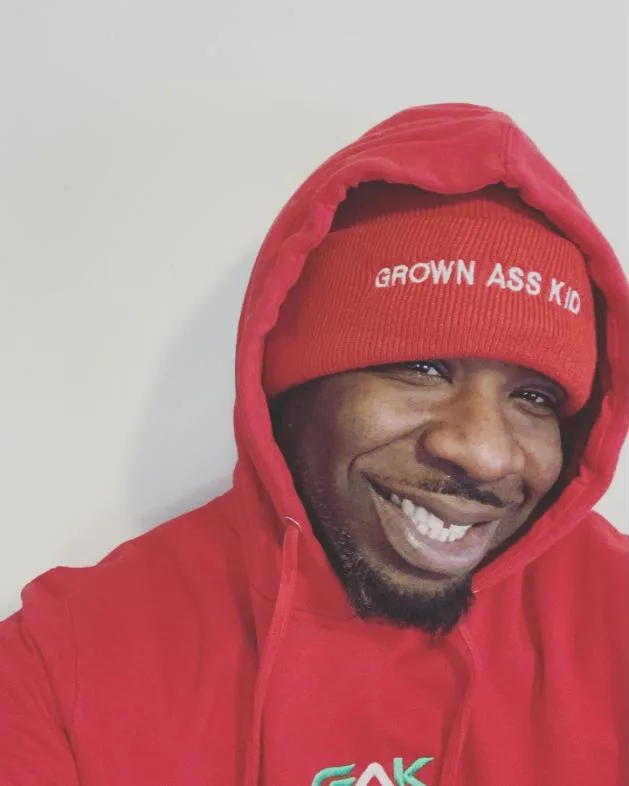 Genaro smiling in a red GAK hoodie and Grown Ass Kid beanie